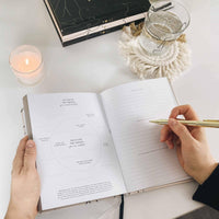 Person holding a pen over an open book on a white surface with a candle and decorative items.