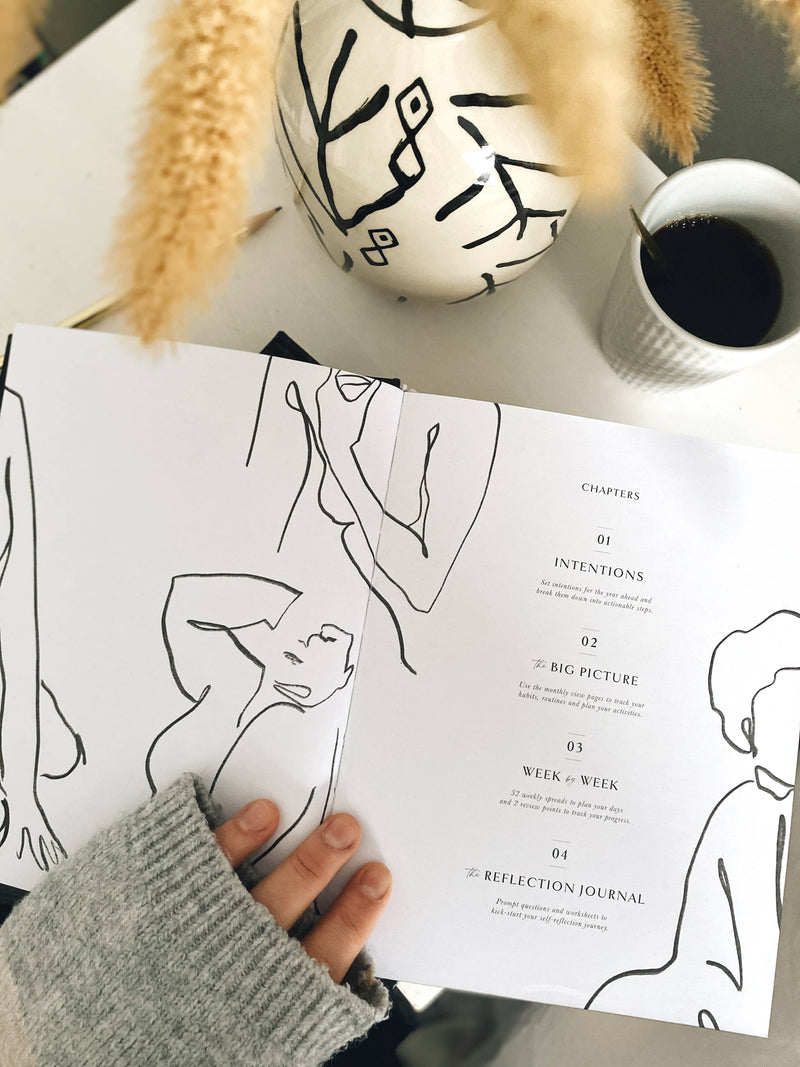 Hand holding a printed sheet with line art and text on a table with a coffee cup and decorative items.