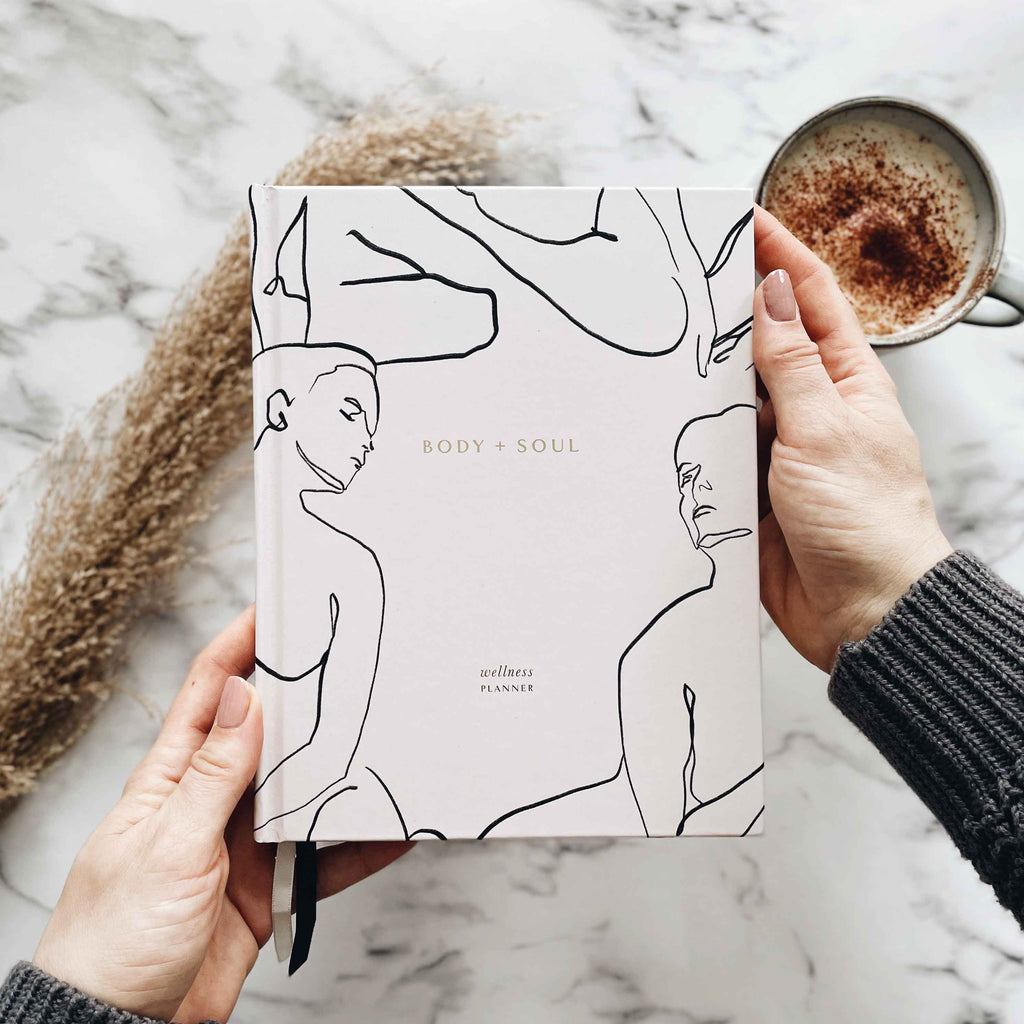 Person holding a planner with line art design on a marble surface with a cup of coffee.