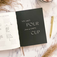 Black notebook with 'You can't pour from an empty cup' text on a marble surface with a journal and pen.