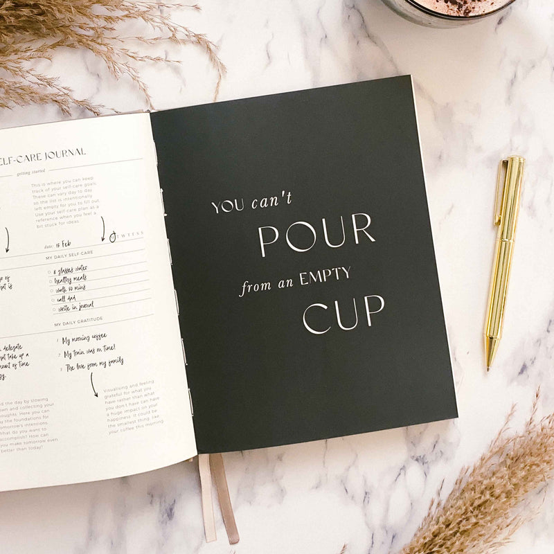 Black notebook with 'You can't pour from an empty cup' text on a marble surface with a journal and pen.