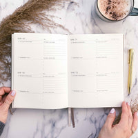 Open planner on a marble surface with a cup of coffee and decorative elements.
