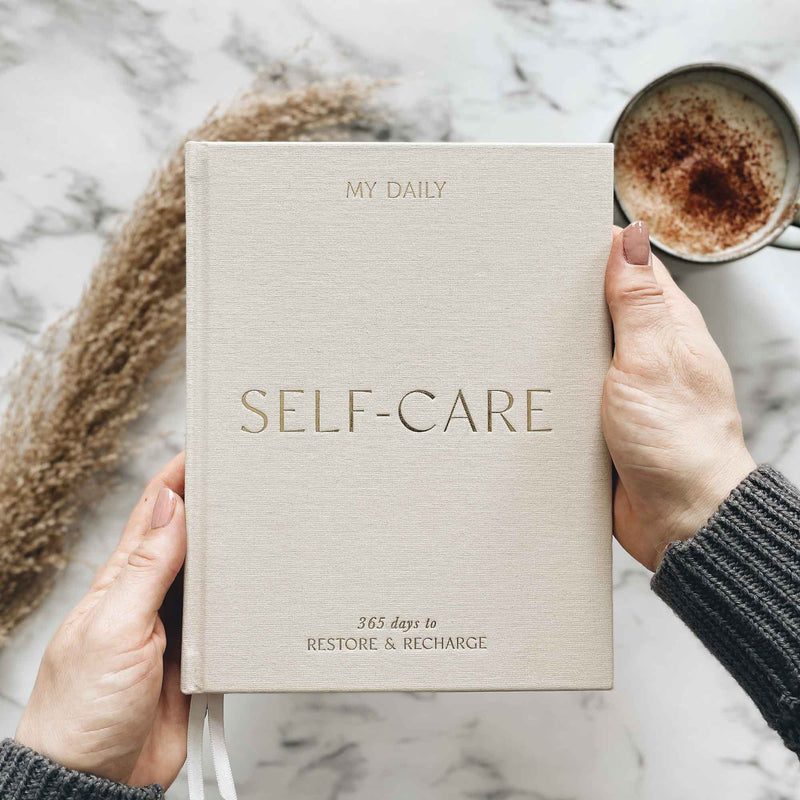 Book titled 'My Daily Self-Care' held by hands with a cup of coffee in the background.