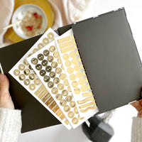 Sheet of decorative stickers with gold and white designs held open by a person.