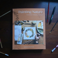 Book titled 'Painting Nature' by Gemma Koomen on a wooden surface with art supplies.