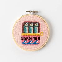 Cross-stitched design of sardines in a can on a pink background within a wooden hoop.