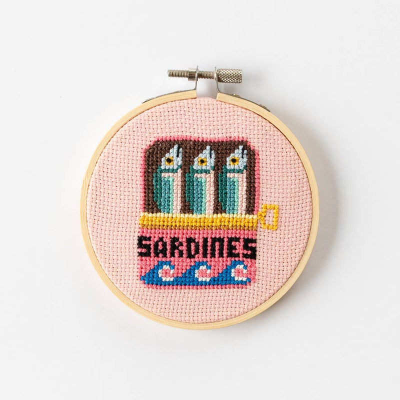 Cross-stitched design of sardines in a can on a pink background within a wooden hoop.