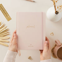 Pink planner held by hands on a light surface with decorative elements