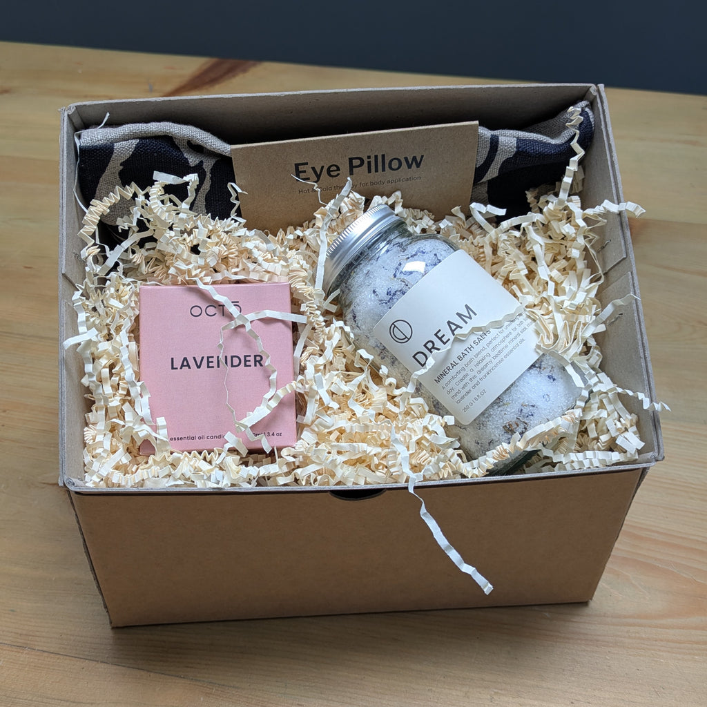 Gift box with bath products including a jar labeled 'Dream', eye pillow, and lavender item on a wooden surface.