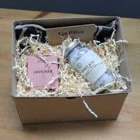 Gift box with bath products including a jar labeled 'Dream', eye pillow, and lavender item on a wooden surface.